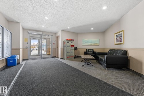 The property entrance features a carpeted floor, glass double doors, recessed lighting, and a seating area with a black sofa - 208 6710 158 Avenue, Edmonton, AB - Indoor Photo Showing Other Room