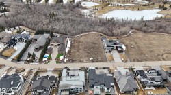 Aerial view of the property and its surrounding area, featuring a paved street, established homes, and undeveloped land bordering a treed area with visible frozen ponds - 