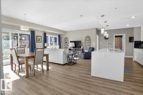 This open-concept living space features light wood-style flooring, a dining area with a modern chandelier, and a kitchen island with pendant lighting - 214 528 Griesbach Parade Parade, Edmonton, AB - Indoor
