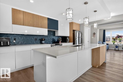Contemporary kitchen featuring white cabinetry, wood-tone upper cabinets, a textured blue backsplash, and a large island with a sink and pendant lighting - 214 528 Griesbach Parade Parade, Edmonton, AB - Indoor Photo Showing Kitchen With Upgraded Kitchen
