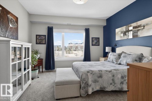 The property features a spacious room with a large window providing views of the surrounding area, complemented by a light-colored carpet, and a white display cabinet with glass doors - 214 528 Griesbach Parade Parade, Edmonton, AB - Indoor Photo Showing Bedroom