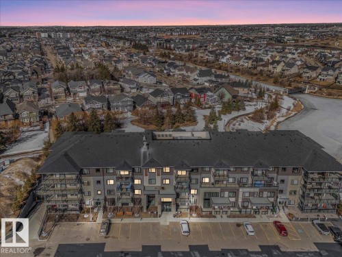 Overhead property view featuring a large building with a dark roof and balconies, a parking area, and a surrounding neighbourhood with residential buildings and a body of water - 214 528 Griesbach Parade Parade, Edmonton, AB - Outdoor With View