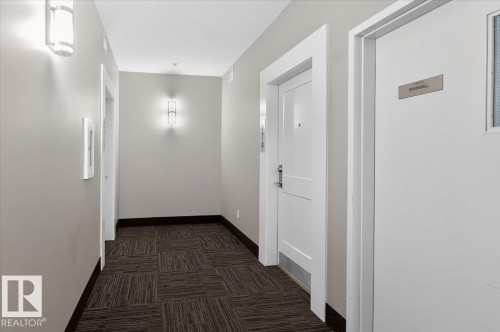 The property features a hallway with patterned carpet flooring, light gray walls, and wall-mounted light fixtures - 214 528 Griesbach Parade Parade, Edmonton, AB - Indoor Photo Showing Other Room