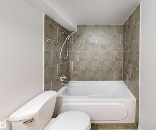 The bathroom features a white bathtub with a shower, tiled walls in a neutral tone with a decorative accent band, and a white toilet - 303 8215 83 Avenue, Edmonton, AB - Indoor Photo Showing Bathroom