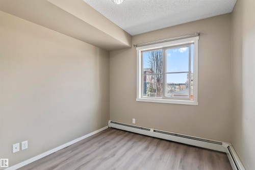 This room features light-colored walls, wood-style flooring, and a large window providing natural light - 214 5816 Mullen Place, Edmonton, AB - Indoor Photo Showing Other Room