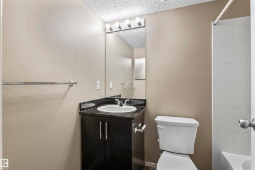The bathroom features a vanity with a dark countertop and an integrated sink, a large mirror with overhead lighting, and a white toilet - 214 5816 Mullen Place, Edmonton, AB - Indoor Photo Showing Bathroom