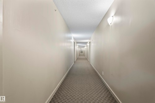 The property features a well-lit corridor with neutral-toned walls, overhead lighting, and wall-mounted sconces providing ample illumination - 214 5816 Mullen Place, Edmonton, AB - Indoor Photo Showing Other Room