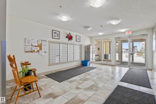 The property features a bright common area with tiled flooring, wall-mounted mailboxes, and an exit door with clear glass panels - 214 5816 Mullen Place, Edmonton, AB - Indoor Photo Showing Other Room