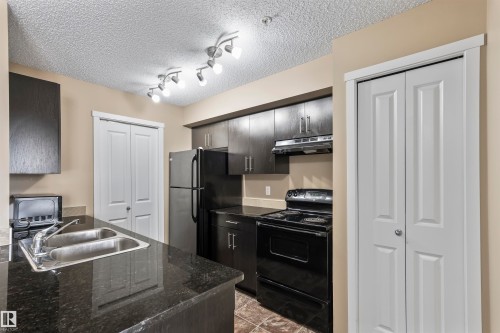 The kitchen features dark cabinetry, a dual basin sink, and a black electric range with an overhead range hood - 214 5816 Mullen Place, Edmonton, AB - Indoor Photo Showing Kitchen With Double Sink