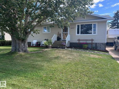 Exterior view of the property featuring light-colored siding, a front yard with a mature tree, and a paved driveway - 4515 54 Ave, Tofield, AB - Outdoor