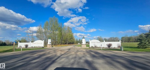 Entryway featuring a paved road, white stone-clad pillars, and surrounding green spaces with mature trees - #1 Sunset Harbour, Rural Wetaskiwin County, AB 
