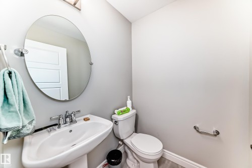 The bathroom features a white pedestal sink with a chrome faucet, an oval mirror, and a toilet - 7822 Koruluk Link Link, Edmonton, AB - Indoor Photo Showing Bathroom