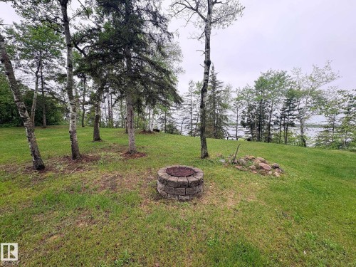 Expansive green space featuring a stone fire pit, mature deciduous trees, evergreen trees, and a distant body of water - #4 Kingbird Road, Rural Bonnyville M.D., AB 