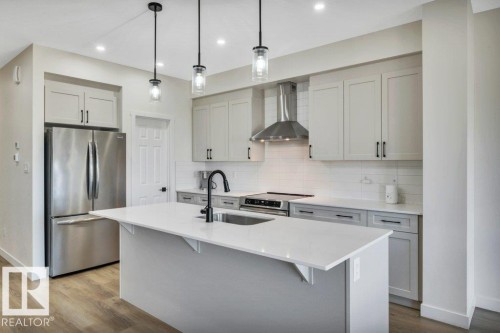 The kitchen features light-toned cabinetry, a stainless steel refrigerator, and a kitchen island with a sink and pendant lighting - 13007 213 Street, Edmonton, AB - Indoor Photo Showing Kitchen With Upgraded Kitchen
