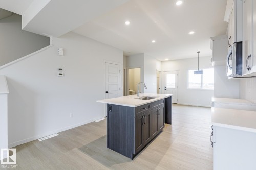 The kitchen features a central island with a sink and dark cabinetry, complemented by light-colored countertops and overhead lighting - 3828 38 Street E, Beaumont, AB - Indoor Photo Showing Kitchen