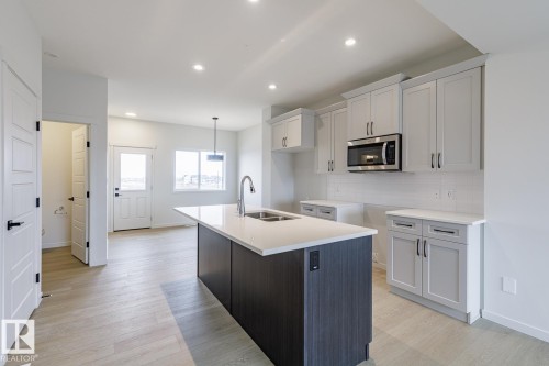 The kitchen features light-toned cabinetry, stainless steel microwave, and an island with a sink and a contemporary pendant light fixture - 3828 38 Street E, Beaumont, AB - Indoor Photo Showing Kitchen With Double Sink