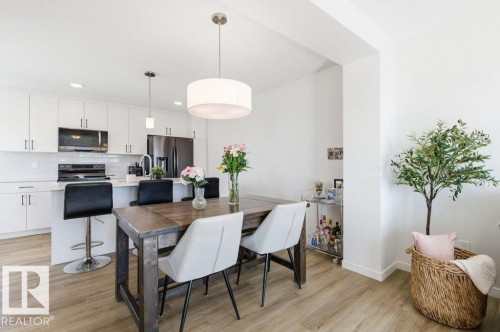 Kitchen and dining area featuring light-toned flooring, white cabinetry, a kitchen island with seating, and a large drum pendant light fixture over the dining table - 49 850 Secord Boulevard, Edmonton, AB - Indoor Photo Showing Other Room