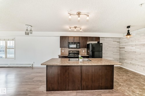 The kitchen features dark wood cabinetry, a central island with a countertop and dual sinks, and black appliances - 301 103 Ambleside Drive, Edmonton, AB - Indoor Photo Showing Kitchen