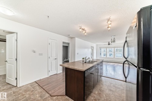 The kitchen features a functional island with a double sink and dark cabinetry, contrasting with the light-toned floor tiles - 301 103 Ambleside Drive, Edmonton, AB - Indoor Photo Showing Kitchen With Double Sink