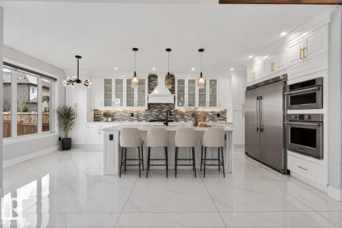 Gourmet kitchen featuring a substantial central island with seating, white shaker cabinetry, stainless steel appliances, and large format polished tile flooring - 707 180 Street, Edmonton, AB - Indoor Photo Showing Kitchen With Upgraded Kitchen