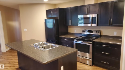 The kitchen features dark cabinetry, stainless steel appliances, and an island with a double sink - 2103 1601 8 Avenue, Cold Lake, AB - Indoor Photo Showing Kitchen With Double Sink