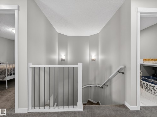 The property features a staircase with a white railing and dark balusters, illuminated by two wall-mounted sconces - 3210 Chernowski Way, Edmonton, AB - Indoor Photo Showing Other Room