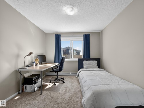 This room features neutral-toned walls, carpeted flooring, and a window with blue curtains, providing natural light - 124 148 Ebbers Boulevard, Edmonton, AB - Indoor