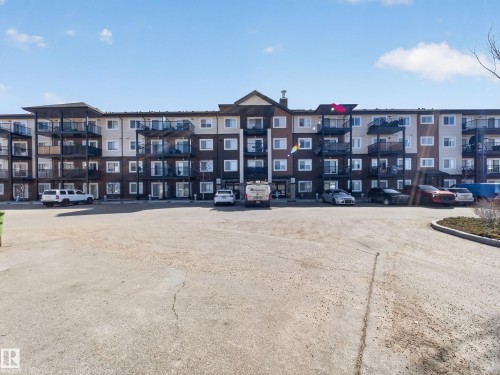 The property features a multi-story building with a brown and beige facade, individual balconies, and a paved parking area - 124 148 Ebbers Boulevard, Edmonton, AB - Outdoor With Facade