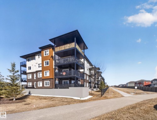 The property features a multi-story building with balconies, a paved pathway, and a grassy area with some evergreen trees - 124 148 Ebbers Boulevard, Edmonton, AB - Outdoor