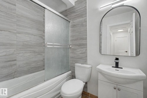 Bathroom featuring a bathtub with sliding frosted glass doors and gray stone-patterned tile, a toilet, and a vanity with a white basin sink, black faucet, and an arched mirror with overhead lighting - 186 Centennial Crest, Edmonton, AB - Indoor Photo Showing Bathroom