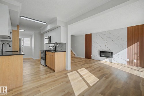 This property features an inviting living space with light-toned flooring, a sleek electric fireplace, and a distinctive accent wall with white marble-patterned paneling and vertical wood panels - 186 Centennial Crest, Edmonton, AB - Indoor Photo Showing Kitchen With Fireplace