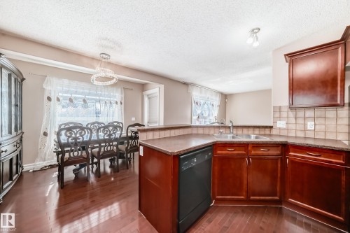The kitchen features dark wood cabinetry, a double basin sink, and a tiled backsplash, with an adjacent dining area showcasing hardwood floors and a decorative light fixture - 11415 167A Avenue, Edmonton, AB - Indoor Photo Showing Kitchen With Double Sink