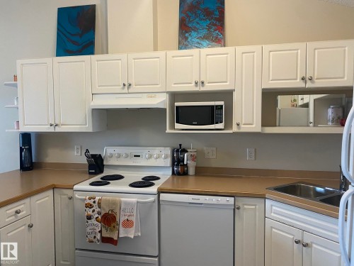 The kitchen features white cabinetry, a white electric range, a microwave, and a dishwasher, complemented by a stainless steel double basin sink - 336 9525 162 Avenue, Edmonton, AB - Indoor Photo Showing Kitchen With Double Sink