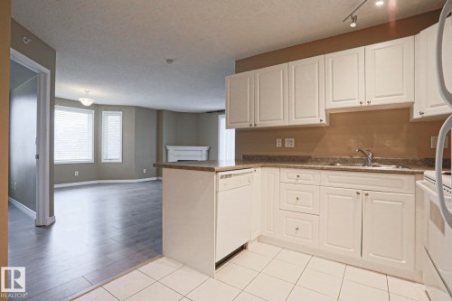 Kitchen featuring white cabinetry, a sink, and a dishwasher, with views of the living area showcasing a fireplace and large windows - 201 10909 103 Avenue Nw, Edmonton, AB - Indoor Photo Showing Kitchen With Double Sink