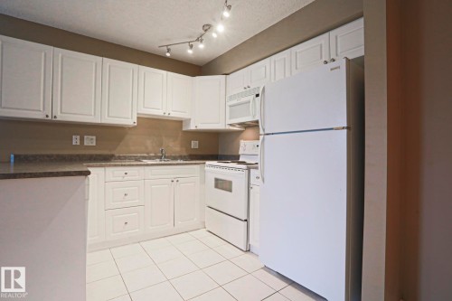 The kitchen features white cabinetry, track lighting, a microwave, and a refrigerator, complemented by a tiled floor - 201 10909 103 Avenue Nw, Edmonton, AB - Indoor Photo Showing Kitchen