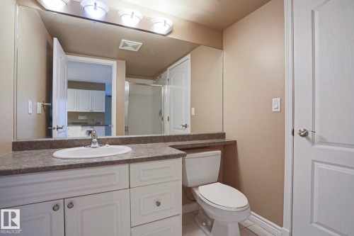 Bathroom featuring a vanity with white cabinetry, a countertop with an integrated sink, a toilet, and a mirror with overhead lighting - 201 10909 103 Avenue Nw, Edmonton, AB - Indoor Photo Showing Bathroom