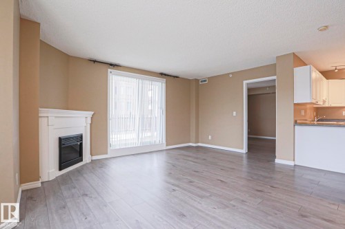The living space features light-colored laminate flooring, a white fireplace with an electric insert, and a large window with sheer white blinds - 201 10909 103 Avenue Nw, Edmonton, AB - Indoor With Fireplace