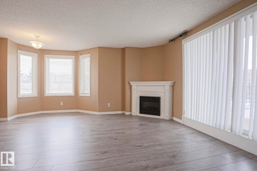 Spacious living area featuring light-colored flooring, a decorative corner fireplace with a white mantel, and a bay window with three window panes - 201 10909 103 Avenue Nw, Edmonton, AB - Indoor Photo Showing Living Room With Fireplace