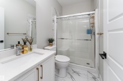 The bathroom features a modern vanity with a white countertop and gold-toned hardware, a spacious shower with a clear glass enclosure, and white marble-patterned floor tiles - 