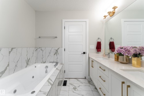 Bathroom featuring a jetted tub with marble-look tile surround and a double vanity with white cabinetry, gold hardware, and a large mirror - 993 Berg Pl, Leduc, AB - Indoor Photo Showing Bathroom