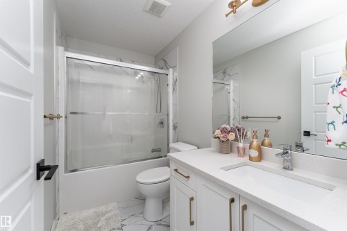 The bathroom features a vanity with a white countertop and gold-toned hardware, a tub with a sliding glass door, and white tiled walls - 993 Berg Pl, Leduc, AB - Indoor Photo Showing Bathroom