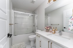 The bathroom features a vanity with a white countertop and gold-toned hardware, a tub with a sliding glass door, and white tiled walls - 