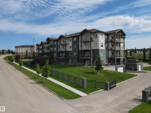 The property features a multi-story building with balconies, a well-maintained lawn, and a paved driveway, all enclosed by a black metal fence with stone pillars - 304 5201 Brougham Drive, Drayton Valley, AB - Outdoor With Balcony With Facade