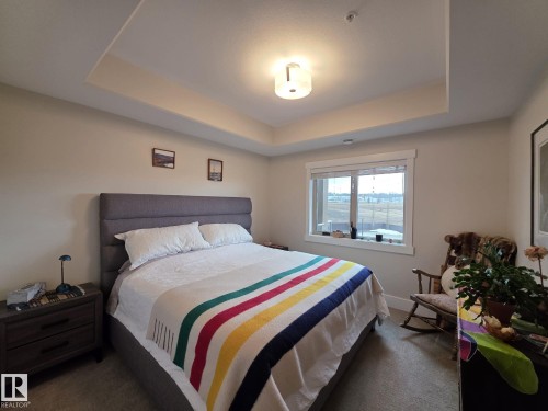 This bedroom features a tray ceiling, a window with views, and a wall-mounted light fixture - 304 5201 Brougham Drive, Drayton Valley, AB - Indoor Photo Showing Bedroom