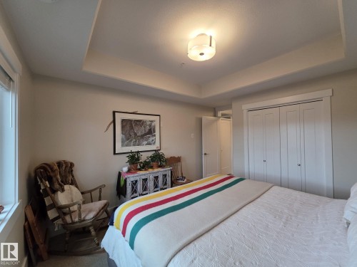 The room features a tray ceiling with a central light fixture, a window with blinds, and white bi-fold closet doors - 304 5201 Brougham Drive, Drayton Valley, AB - Indoor Photo Showing Bedroom