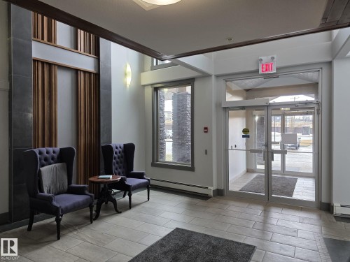 Welcoming entry area with light-toned tiled flooring, featuring two dark upholstered armchairs, a round side table, and large windows providing natural light - 304 5201 Brougham Drive, Drayton Valley, AB - Indoor Photo Showing Other Room