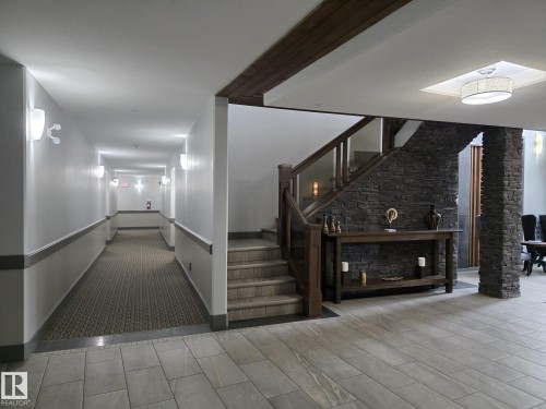 This property features a well-lit hallway with wall sconces and patterned carpeting, along with a staircase featuring a wooden handrail and glass panels - 304 5201 Brougham Drive, Drayton Valley, AB - Indoor Photo Showing Other Room