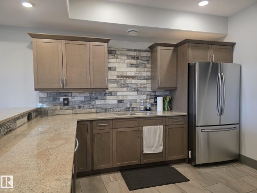 The property includes a kitchen space featuring granite countertops, wood cabinetry, a stainless steel refrigerator, and a tiled backsplash - 304 5201 Brougham Drive, Drayton Valley, AB - Indoor Photo Showing Kitchen