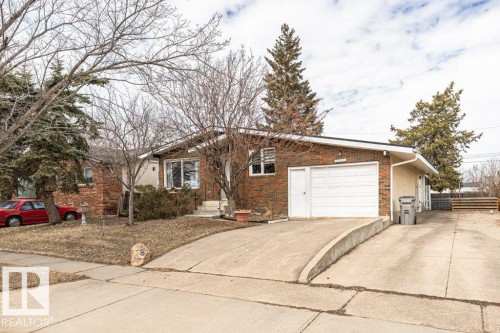 The property features a brick exterior with a white garage door, a concrete driveway, and established trees in the front yard - 8 Duke Drive, Lamont, AB - Outdoor