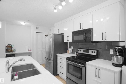 The kitchen features white cabinetry, a double basin stainless steel sink, and a dark subway tile backsplash - 103 3353 16A Avenue, Edmonton, AB - Indoor Photo Showing Kitchen With Double Sink